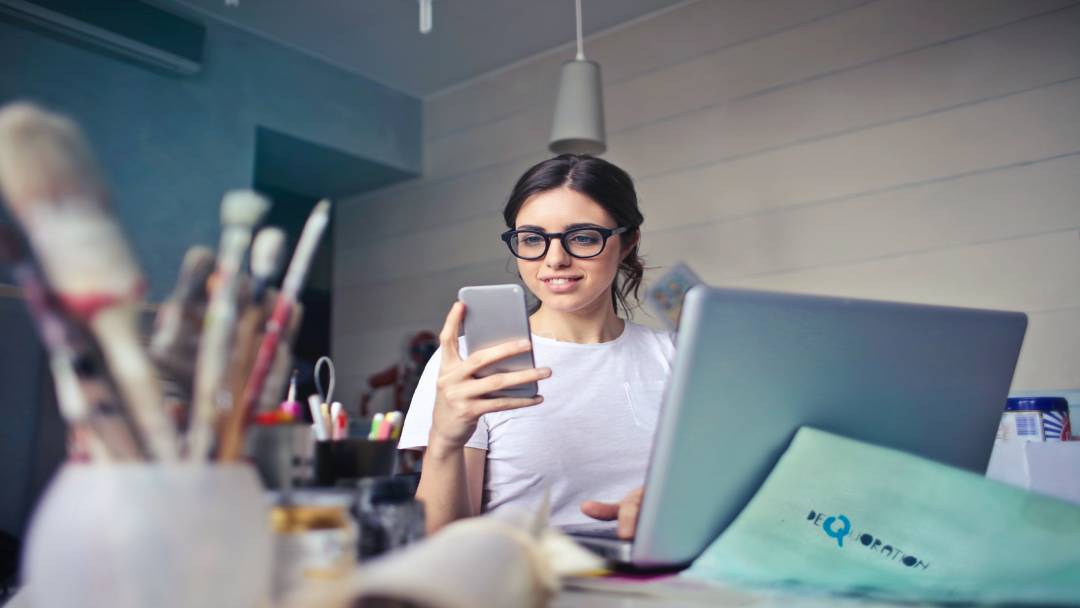 Woman looking at her phone with laptop and brushes nearby